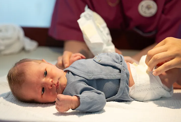 Ein Säugling liegt auf einem Wickeltisch, während eine Person in roter Kleidung eine Windel wechselt. Der Raum ist hell und freundlich gestaltet.