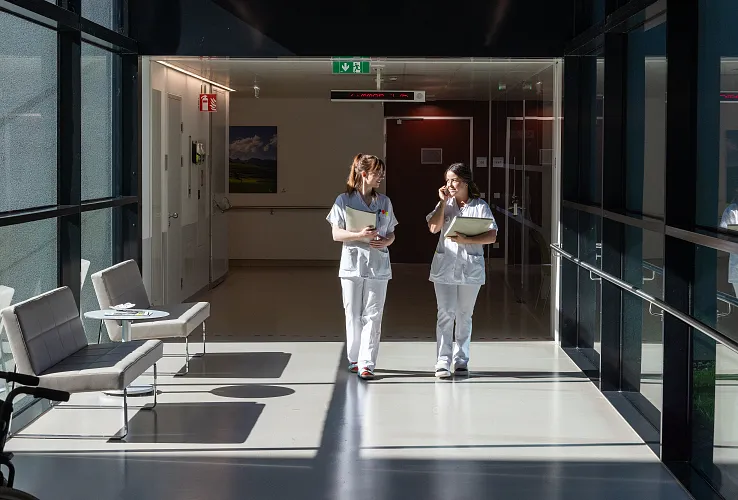 Zwei Krankenhausmitarbeiterinnen in weißer Arbeitskleidung gehen einen hell beleuchteten Flur entlang und unterhalten sich. Stühle stehen seitlich an der Wand.