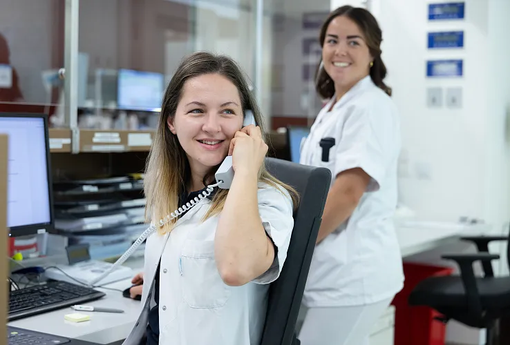 Zwei medizinische Fachkräfte in weißen Arbeitsuniformen arbeiten an einem Empfangsschalter im Krankenhaus. Eine telefoniert, die andere lächelt in die Kamera.