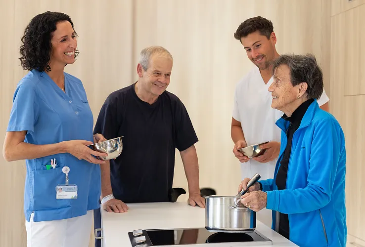 Vier Personen, darunter Pflegepersonal und ältere Menschen, kochen gemeinsam in einer modernen Küche. Sie lachen und halten Küchengeräte.