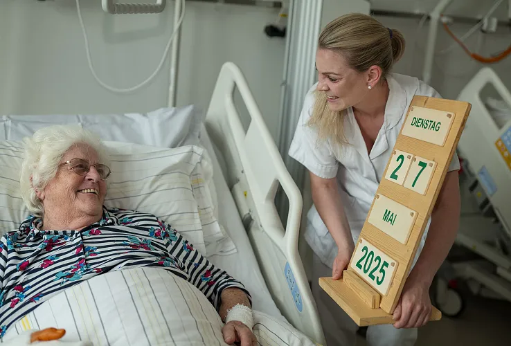 Eine Krankenschwester hält ein Datumsbrett mit dem Datum "27. Mai 2025" neben einer lächelnden älteren Frau im Krankenbett.