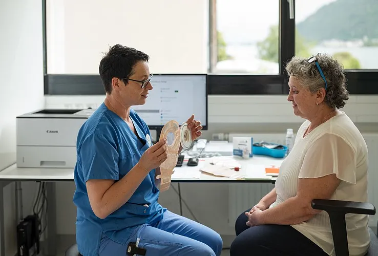 Gesundheitsfachkraft in blauem Kittel zeigt einer älteren Frau medizinisches Hilfsmittel. Beide sitzen in einem hellen Raum mit einem Fenster im Hintergrund.