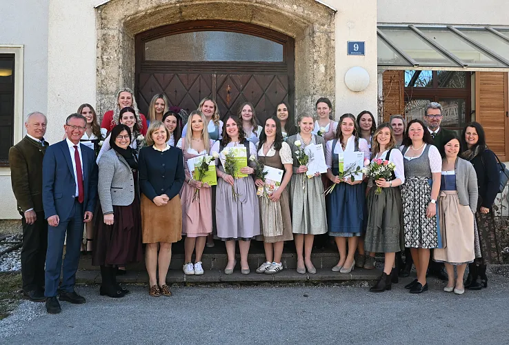 Eine Gruppe von Menschen steht lächelnd vor einem Gebäude. Einige tragen traditionelle Trachten und halten Blumensträuße und Urkunden in den Händen.