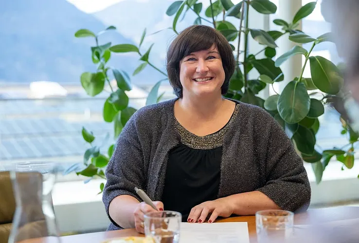 Lächelnde Frau mit dunklem Haar sitzt an einem Tisch, umgeben von Pflanzen. Sie hält einen Stift über einem Dokument. Im Hintergrund sind Fenster und Berge sichtbar.