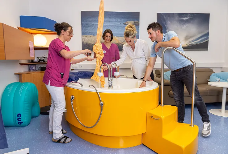 Geburtsvorbereitungskurs in einem modernen Kreißsaal. Eine Gruppe von Menschen um eine gelbe Geburtswanne, begleitet von Hebammen, die Anweisungen geben.