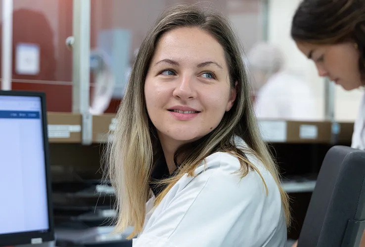 Labormitarbeiter:Innen bei der Arbeit. Eine Frau lächelt, während sie vor einem Computer sitzt. Weitere Person im Hintergrund unscharf erkennbar.