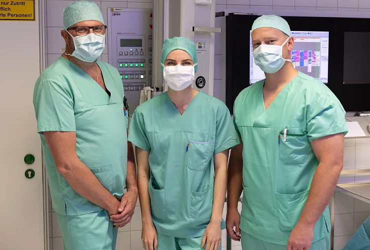 Drei Personen in OP-Kleidung stehen in einem Krankenhausraum. Sie tragen grüne Kittel, Hauben und Masken und blicken in die Kamera.