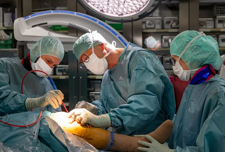Drei Chirurgen in OP-Kleidung führen eine Operation durch. Sie arbeiten konzentriert unter heller Beleuchtung, umgeben von medizinischen Geräten und sterilen Materialien.