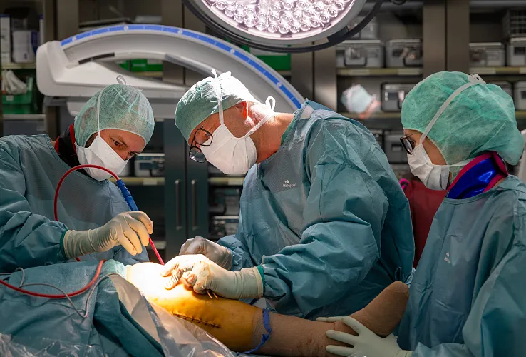 Drei Chirurgen in OP-Kleidung operieren in einem hell ausgeleuchteten Operationssaal. Sie tragen grüne Kittel, Hauben und Masken und arbeiten konzentriert.