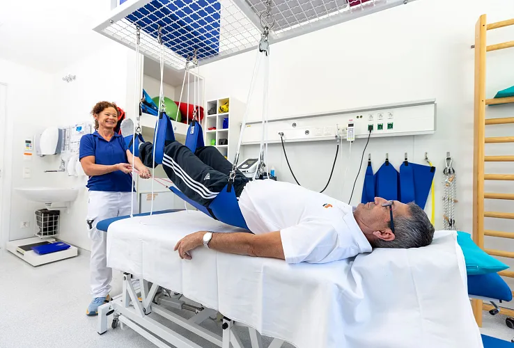 Ein Patient liegt in bequemer Kleidung auf einer Behandlungsliege, während eine Therapeutin eine Übung mit den Beinen des Patienten durchführt.