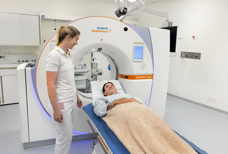 Eine medizinische Szenerie zeigt einen Patienten, der für ein CT-Scan auf einem Tisch liegt. Eine Krankenschwester steht lächelnd daneben. Der Raum ist modern.