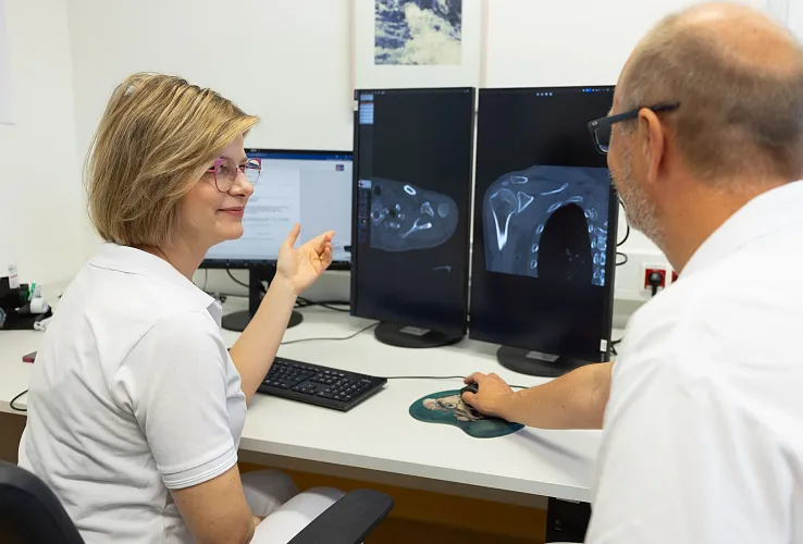Zwei medizinische Fachleute besprechen Röntgenbilder, die auf zwei Monitoren angezeigt werden. Sie tragen weiße Kittel und scheinen in einem Büro zu arbeiten.