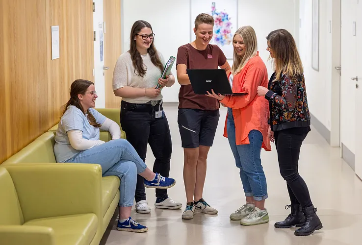 Eine Gruppe von fünf jungen Erwachsenen steht in einem modernen Gang. Vier stehen zusammen und schauen auf einen Laptop, eine Person sitzt auf einem grünen Sofa.
