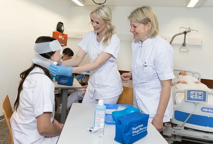 Zwei Krankenschwestern versorgen eine sitzende Patientin mit einem Kopfverband in einem Krankenhauszimmer. Im Hintergrund steht ein Bett mit einem Patienten.