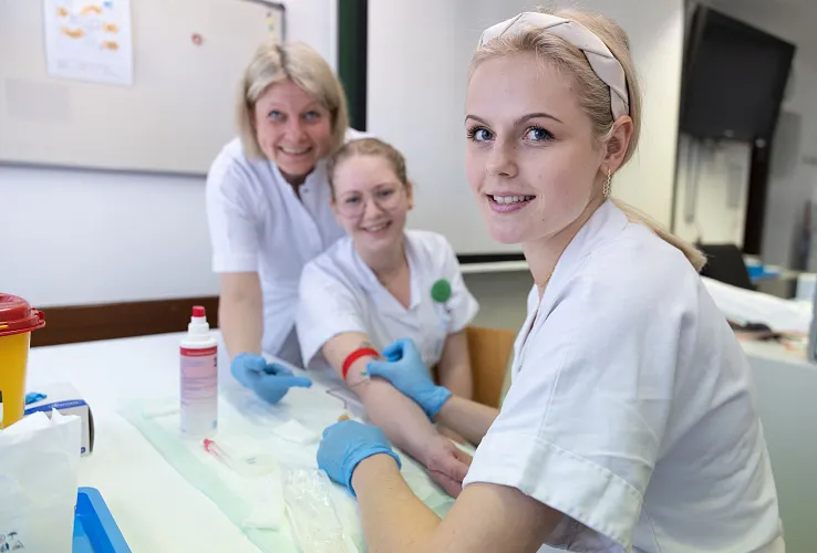Drei Frauen in medizinischer Kleidung sitzen an einem Tisch. Eine führt eine Blutabnahme bei der anderen durch. Alle lächeln freundlich in die Kamera.