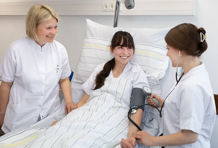 Zwei Ärztinnen betreuen eine lächelnde Patientin im Krankenhausbett. Eine misst den Blutdruck, während die andere die Hand der Patientin hält.
