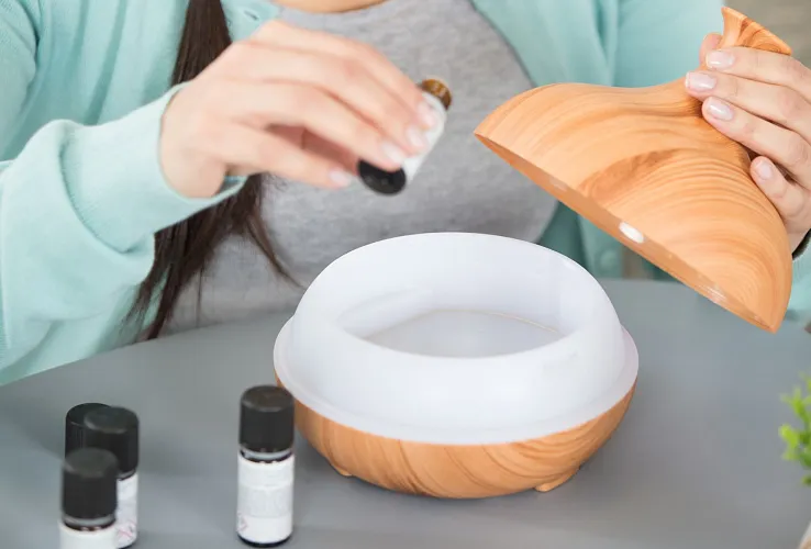 Frau mit langen, geflochtenen Haaren tropft ätherisches Öl in einen Holz-Diffusor. Sie trägt einen türkisfarbenen Cardigan und ein graues T-Shirt.