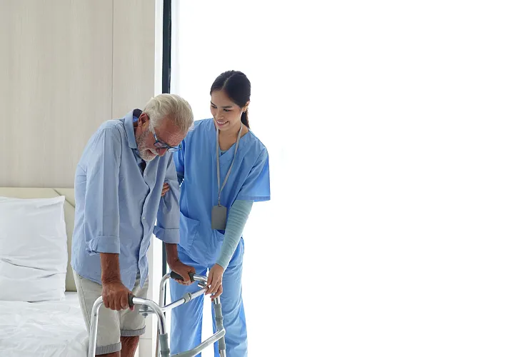 Eine Krankenschwester unterstützt einen älteren Mann mit einem Gehhilfe beim Gehen in einem hellen Raum. Beide wirken konzentriert und vertraut miteinander.