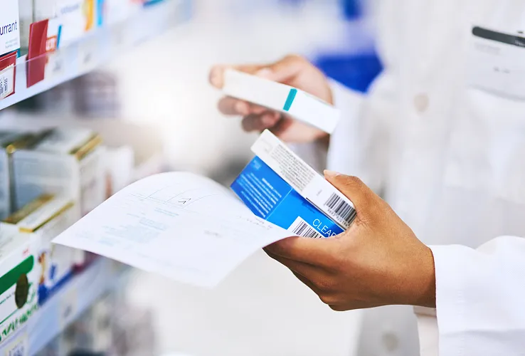 Person in weißem Kittel hält ein Rezept und eine Medikamentenschachtel. Im Hintergrund sind Regale mit anderen Medikamenten zu sehen.