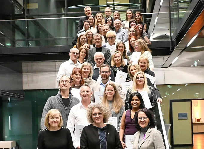 Eine Gruppe von Menschen steht auf einer Treppe in einem modernen Gebäude. Sie lächeln und halten Urkunden oder Dokumente in den Händen.