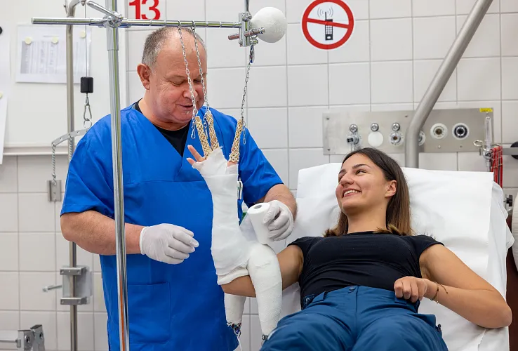 Ein Arzt in blauer Uniform versorgt den Gipsverband einer lächelnden Patientin, die auf einem Krankenhausbett sitzt. Beide scheinen entspannt und zufrieden.