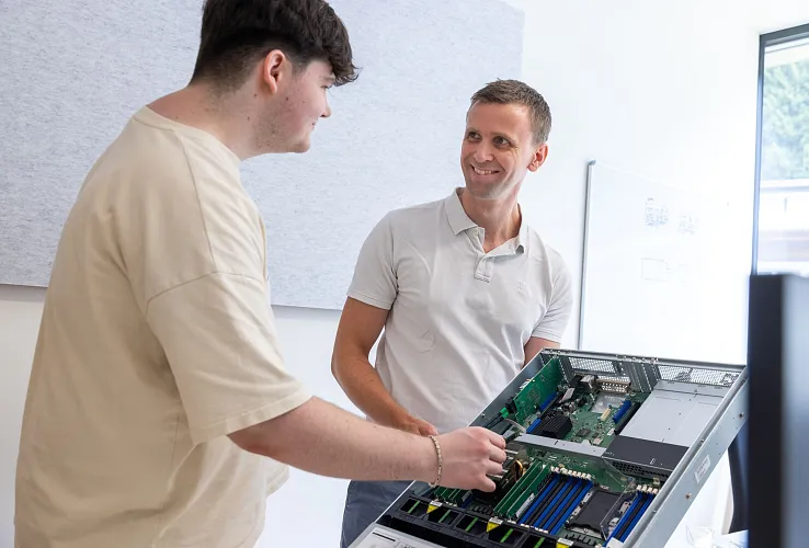 Zwei Männer betrachten und diskutieren über die interne Hardware eines offenen Computer-Servers in einem modern ausgestatteten Büro mit Akustikpanelen.
