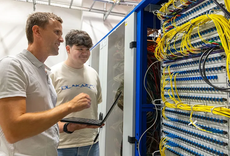 Zwei Männer arbeiten in einem Technikraum an einem offenen Netzwerk-Schrank voller Kabel. Einer hält einen Laptop, beide schauen auf die Verkabelung.
