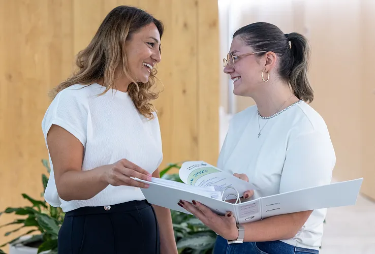 Zwei Frauen in weißen Blusen stehen lächelnd zusammen in einem modern eingerichteten Raum und blättern in einem großen Ordner mit Dokumenten.