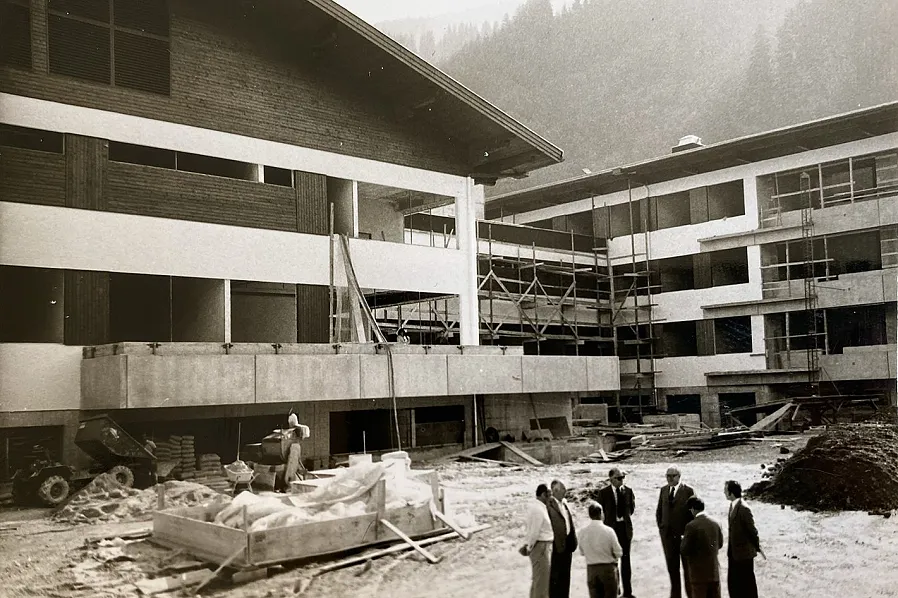 Baustelle eines modernen Gebäudes in den Bergen, umgeben von Bauarbeitern und Ingenieuren, die in einer Gruppe Pläne besprechen; Baumaterialien im Vordergrund.