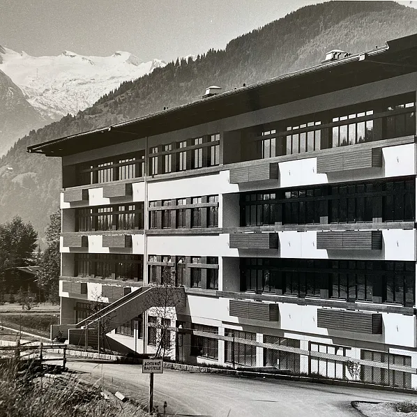 Ein mehrstöckiges, modernes Gebäude mit großen Fenstern vor einer beeindruckenden Bergkulisse. Im Hintergrund erhebt sich ein schneebedeckter Gipfel.