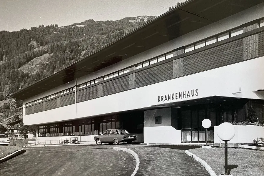 Ein Krankenhausgebäude im 20. Jahrhundert-Stil, umgeben von Bergen. Ein klassisches Auto steht vor dem Eingang. Das Gebäude hat große Fenster.