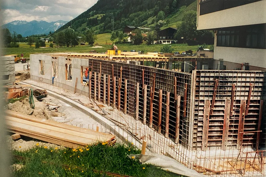 Eine Baustelle mit einer großen Betonwand in einem ländlichen Gebiet, umgeben von Bergen und grünen Wiesen. Bauarbeiter arbeiten an der Konstruktion.