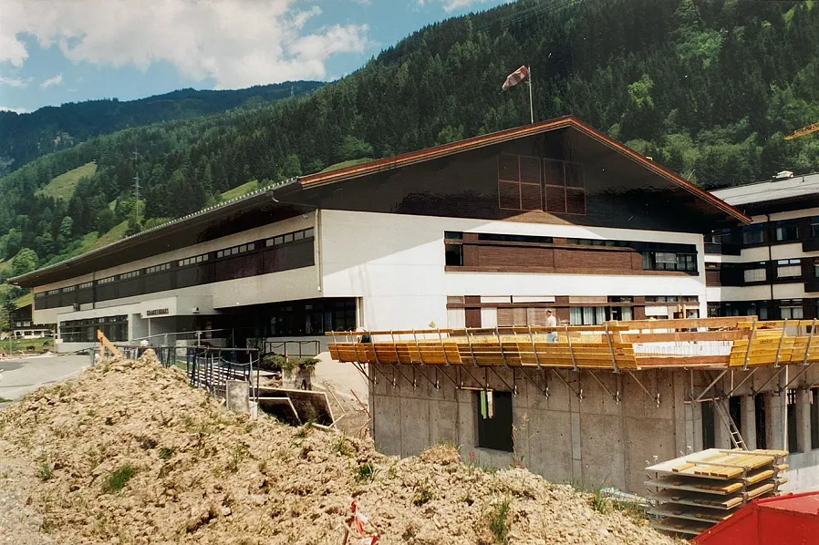 Ein modernes Gebäude mit Holzelementen in einer bergigen Landschaft. Im Vordergrund befindet sich eine Baustelle mit Betonmauern und Holzgerüsten.