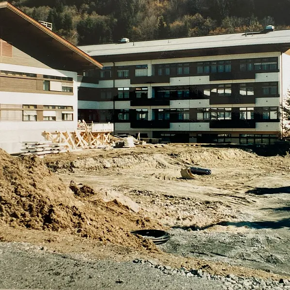 Eine Baustelle vor zwei großen Gebäuden in einer bergigen Landschaft. Ein Bagger arbeitet neben einem Lastwagen auf unebenem, erdigem Gelände.