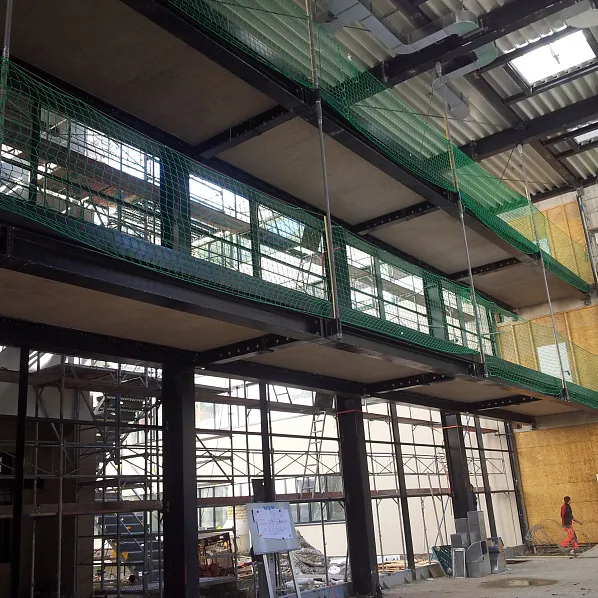 Ein modernes Gebäude im Bau mit Stahlträgern, großen Glasflächen und grünen Schutznetzen. Eine Person geht im Hintergrund, umgeben von Baugerüsten.