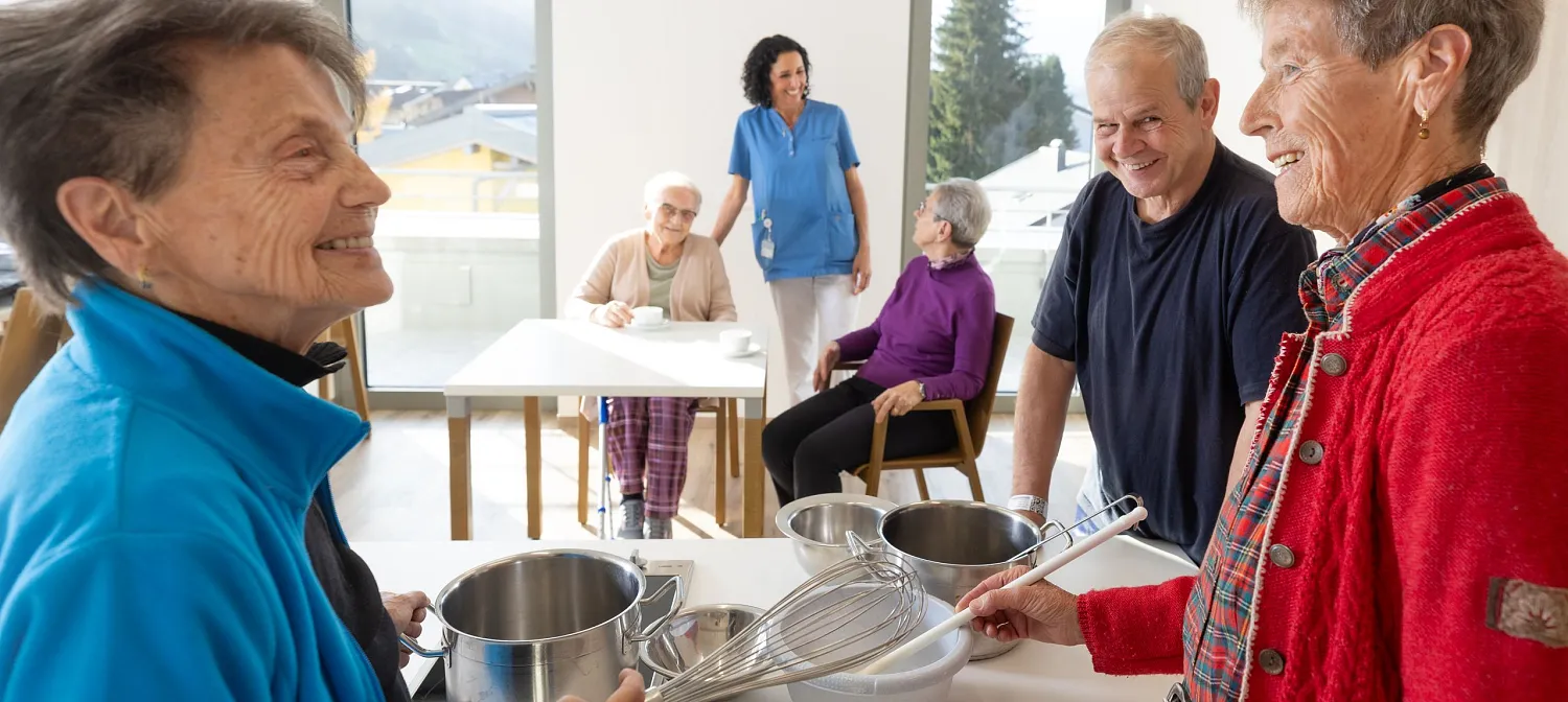 Senior:Innen und Pflegekräfte in gemütlicher Umgebung. Einige halten Küchengeräte, während andere an einem Tisch sitzen und sich unterhalten.