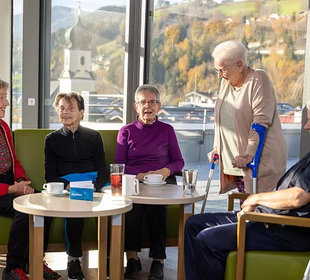 Ältere Erwachsene sitzen in einem hellen Raum mit großen Fenstern, genießen Kaffee und unterhalten sich. Eine Person nutzt Gehhilfen.