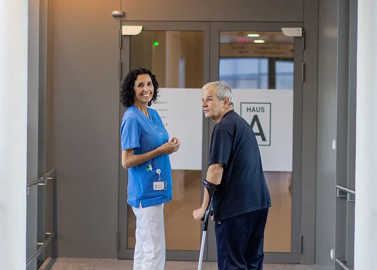 Eine Pflegekraft in blauer Uniform lächelt während sie einem älteren Mann mit Gehstock in einem Krankenhausflur assistiert. Im Hintergrund befindet sich eine Tür mit der Aufschrift 'Haus A'.