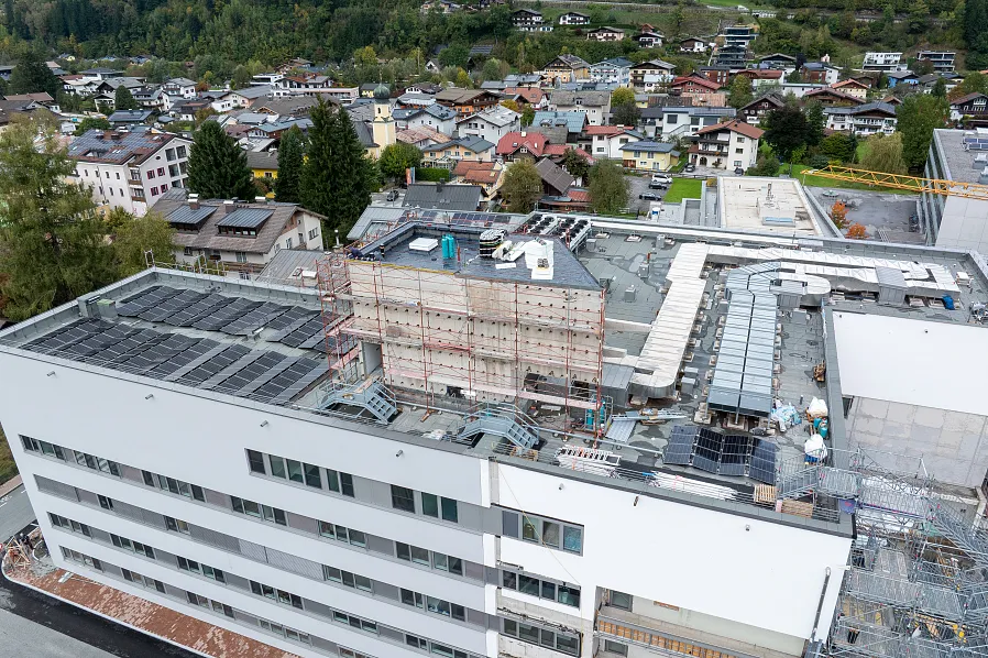 Luftaufnahme eines großen Gebäudekomplexes mit Solarpanelen auf dem Dach, umgeben von Bäumen und einer Wohnsiedlung in einer hügeligen Landschaft.