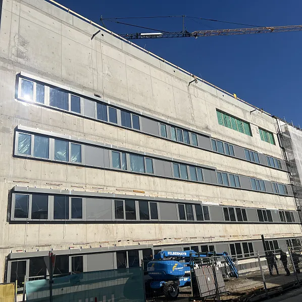 Ein großes, mehrstöckiges Gebäude im Bau, mit Betonwänden und zahlreichen Fenstern. Baugerüste und ein Kran sind sichtbar unter klarem Himmel.
