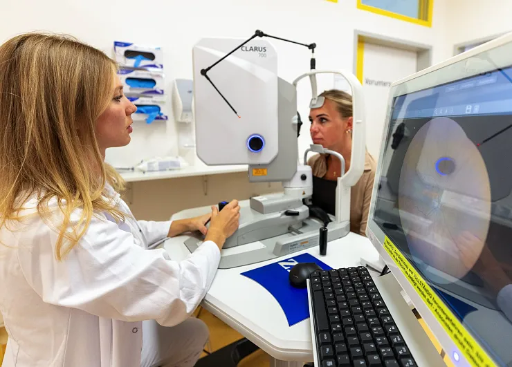 Eine Frau in einem weißen Kittel bedient ein augendiagnostisches Gerät, während eine andere Frau in das Gerät schaut. Auf einem Bildschirm ist ein Augenbild zu sehen.