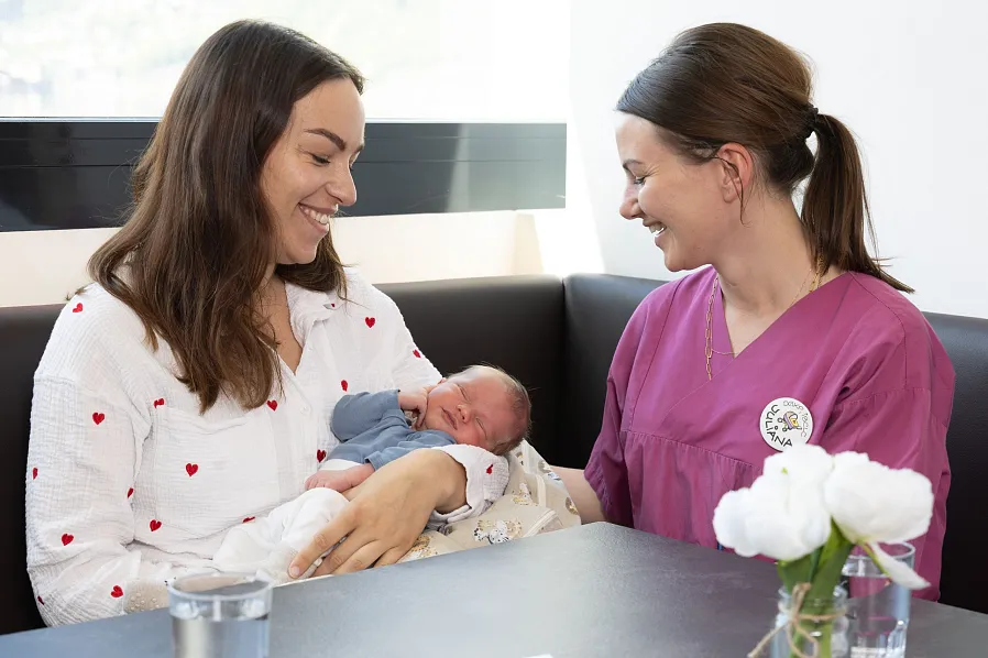 Eine Frau hält ein neugeborenes Baby in einem blauen Strampler, während eine andere Frau in lila Kleidung lächelnd daneben sitzt. Im Vordergrund stehen weiße Blumen.