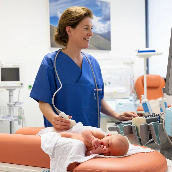 Eine Krankenschwester in blauer Uniform untersucht ein Neugeborenes auf einem orangefarbenen Untersuchungstisch in einer modernen medizinischen Einrichtung.