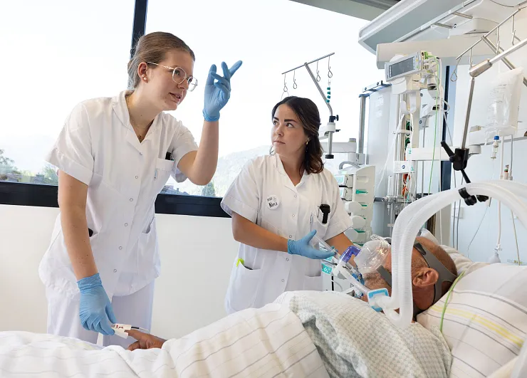 Zwei medizinische Fachkräfte in weißer Arbeitskleidung betreuen einen Patienten im Krankenhausbett. Eine spricht und zeigt gestikulierend mit der Hand.