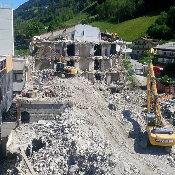 Abriss eines großen Gebäudes mit mehreren Baggern, die Trümmer entfernen. Im Hintergrund eine grüne Hügellandschaft und Berge unter blauem Himmel.