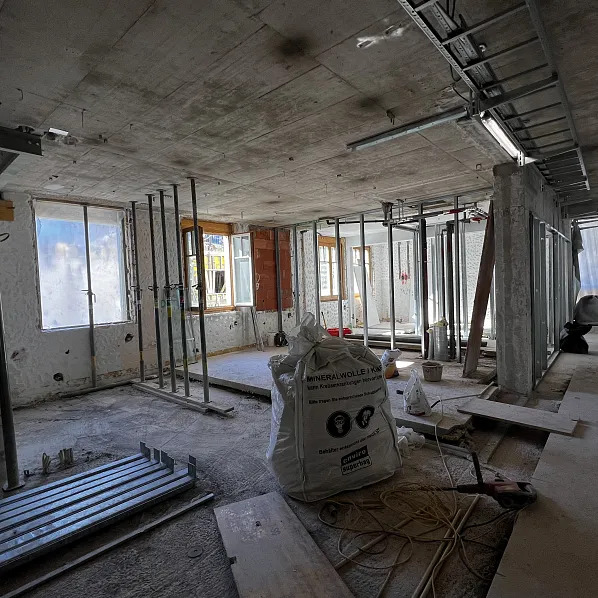 Innenaufnahme einer Baustelle mit sichtbaren Betonwänden und Metallstützen. Ein großer Zementsack liegt am Boden, umgeben von Werkzeugen und Baumaterial.