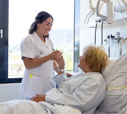 Eine Pflegerin lächelt, während sie die Hand einer älteren, im Krankenhausbett liegenden Patientin hält. Helle Umgebung mit großem Fenster im Hintergrund.