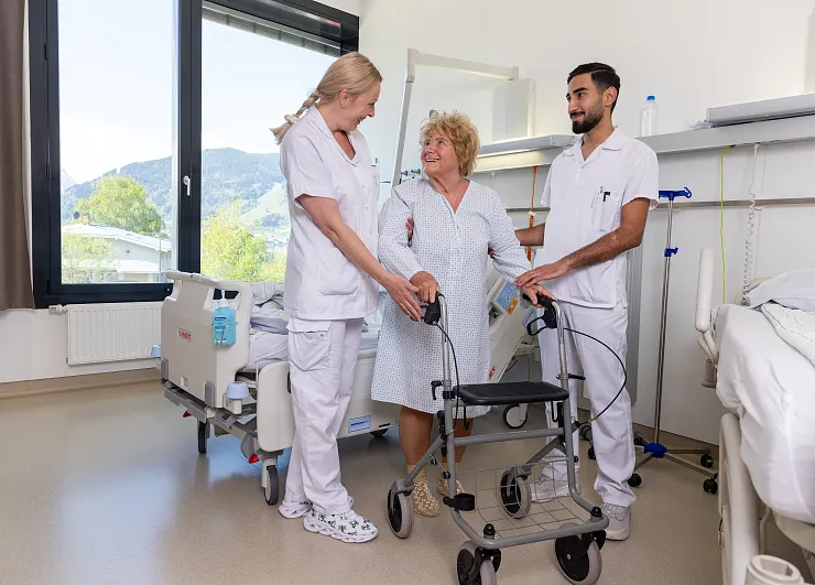 Drei medizinische Fachkräfte in weißen Kitteln stehen in einem Krankenhauszimmer. Sie lächeln und umarmen sich gegenseitig vor einem Rollator.
