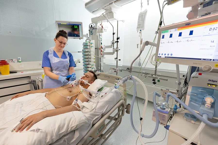 Eine Krankenschw kümmert sich um einen Patienten im Krankenhaus. Der Patient liegt im Bett und ist an medizinische Geräte angeschlossen. Der Raum ist klinisch und hell.