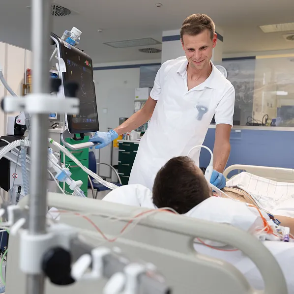 Ein Pfleger in weißer Kleidung untersucht einen Patienten mit einem Ultraschallgerät in einem Krankenhauszimmer. Der Patient liegt im Bett, umgeben von medizinischen Geräten.